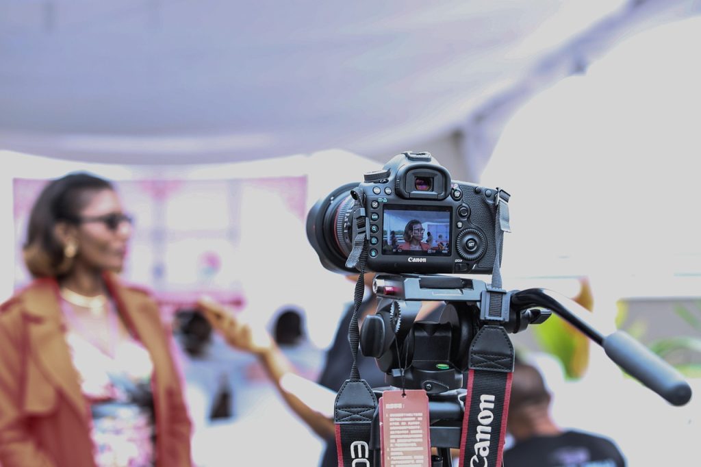 A Canon DSLR camera mounted on a tripod is recording a woman in a red jacket and sunglasses during an interview. The woman appears on the camera’s screen, in focus, while she is blurred in the background. Another person holds a smartphone or microphone toward her, suggesting a media or public event taking place in a tented, well-lit space.