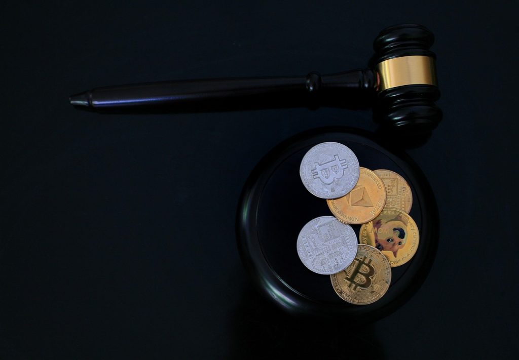 A judge's gavel rests on a circular base next to various cryptocurrency coins, including Bitcoin, Ethereum, and Dogecoin, symbolizing the intersection of legal regulation and digital currencies on a dark background.