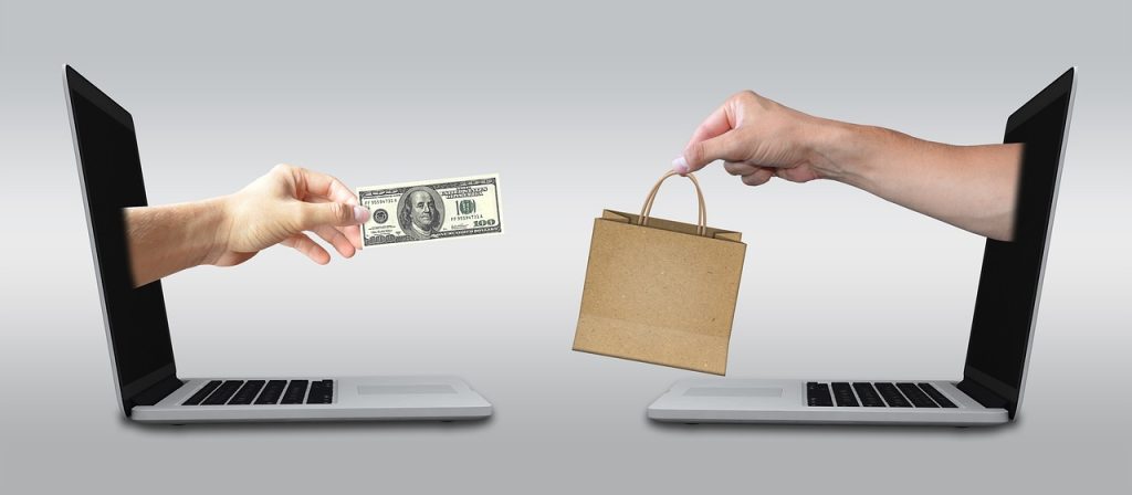 Two laptops face each other, with one hand extending money and the other offering a shopping bag, symbolizing an online purchase transaction.
