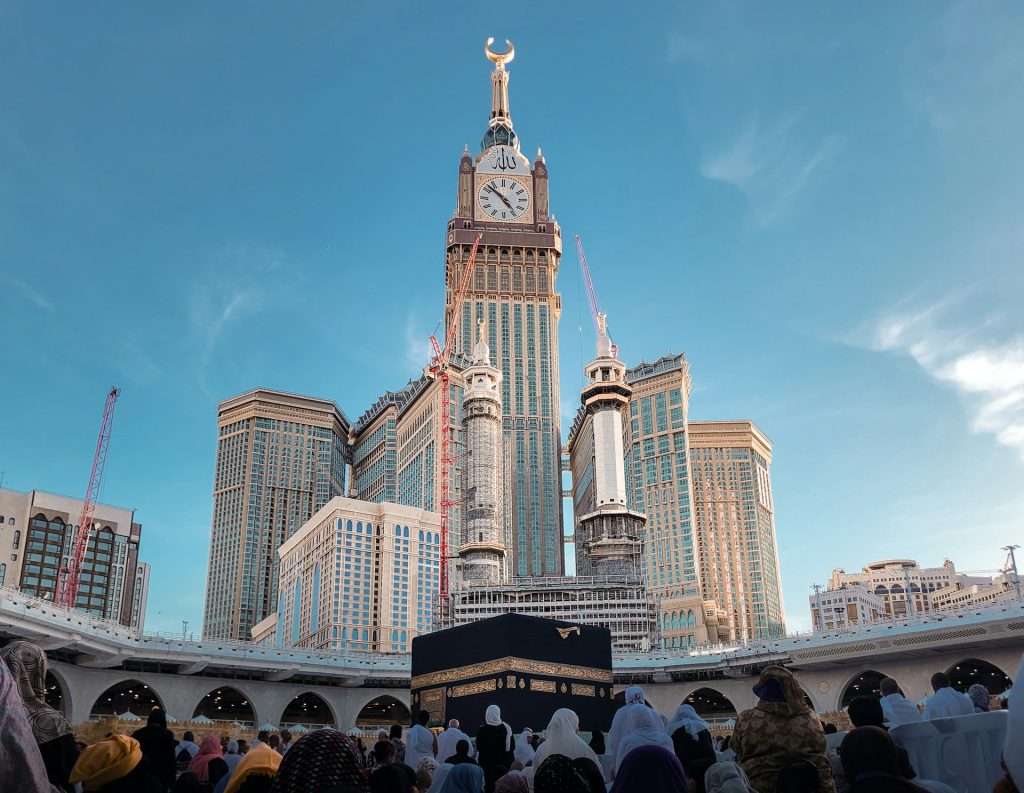 Worshippers gather around the Kaaba at Masjid al-Haram in Mecca, with the Abraj Al-Bait Clock Tower rising in the background under a clear blue sky.