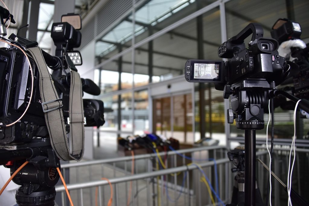  Professional video cameras set up on tripods point toward a row of microphones arranged for a press conference outside a modern glass building. The cameras are positioned behind a metal barricade, and multiple colored cables run along the ground. The setting appears to be prepared for a media event or official statement.