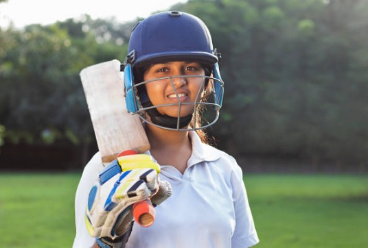 "Female cricketer in helmet and uniform holding a bat, standing on a green field."
