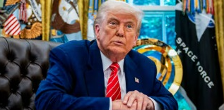 A man in a blue suit and red tie sits at a desk in a formal setting, with U.S. and Space Force flags in the background.