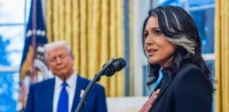 The image shows a woman with long dark hair and a distinctive streak of white speaking at a microphone, appearing confident and composed. She is wearing a dark outfit and has one hand placed over her chest, suggesting a solemn or heartfelt moment. In the background, a man with light-colored hair in a blue suit, red tie, and white shirt is slightly out of focus, standing in front of golden curtains and the U.S. flag, indicating that the scene likely takes place in a formal or governmental setting, such as the Oval Office.