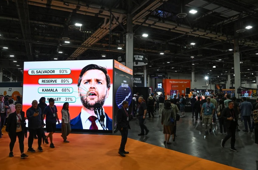 A large digital display at a convention shows a man speaking at a podium, with poll results beside him, while attendees walk around the busy event floor.