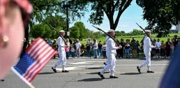 The image depicts a parade scene. In the foreground, a person is holding a small American flag. In the mid-ground, three uniformed military personnel in white dress uniforms are marching with rifles. A crowd of spectators stands along the sidewalk behind them, watching the procession. The scene is set outdoors on a sunny day, with trees providing shade and a clear blue sky in the background. This appears to be a patriotic or ceremonial event, possibly related to Memorial Day or Independence Day.