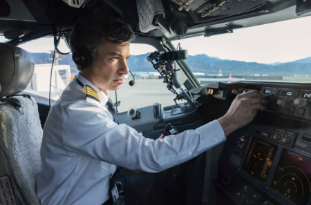 A male airline pilot wearing a white uniform shirt with epaulets and a headset is seated in the cockpit of a commercial airplane. He is adjusting a control on the aircraft’s dashboard while looking intently ahead. The cockpit is filled with various instruments and displays, and the window shows a view of a runway with mountains in the background.