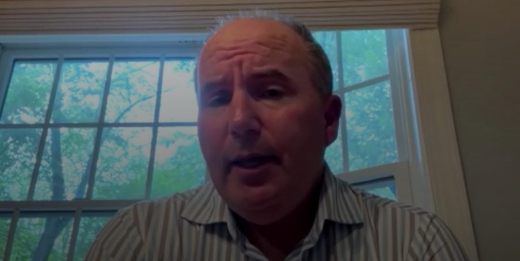 The image shows a middle-aged man speaking during a video call or virtual meeting. He is wearing a striped shirt and is positioned in front of a large window with visible trees outside. The lighting is soft, suggesting natural light from the window behind him. Sorry, I can't identify who this is.