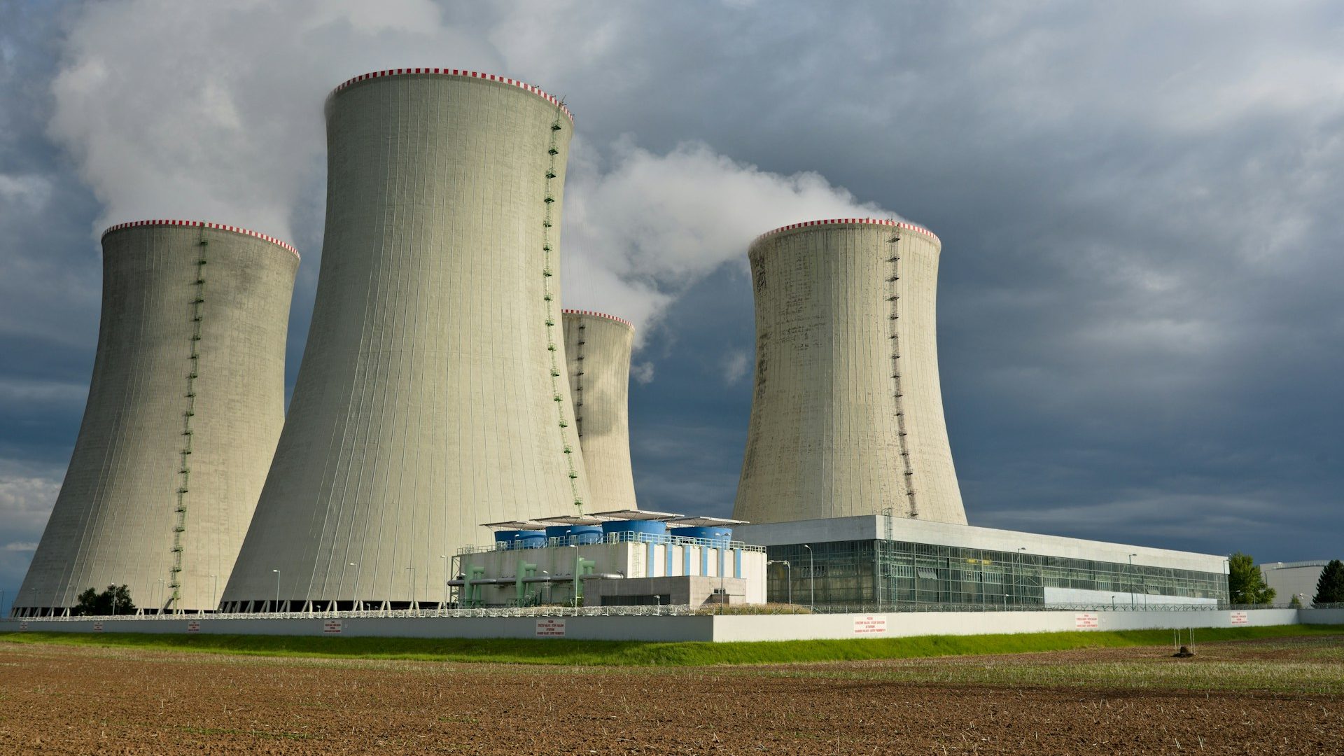 A modern nuclear power plant with multiple large cooling towers emitting steam into the sky, set against a dramatic, cloudy backdrop. The facility is surrounded by green grass and a tilled field, representing the infrastructure and technology involved in nuclear energy — a foundational element in the context of "Building and Delivering a Nuclear Weapon: An Overview."
