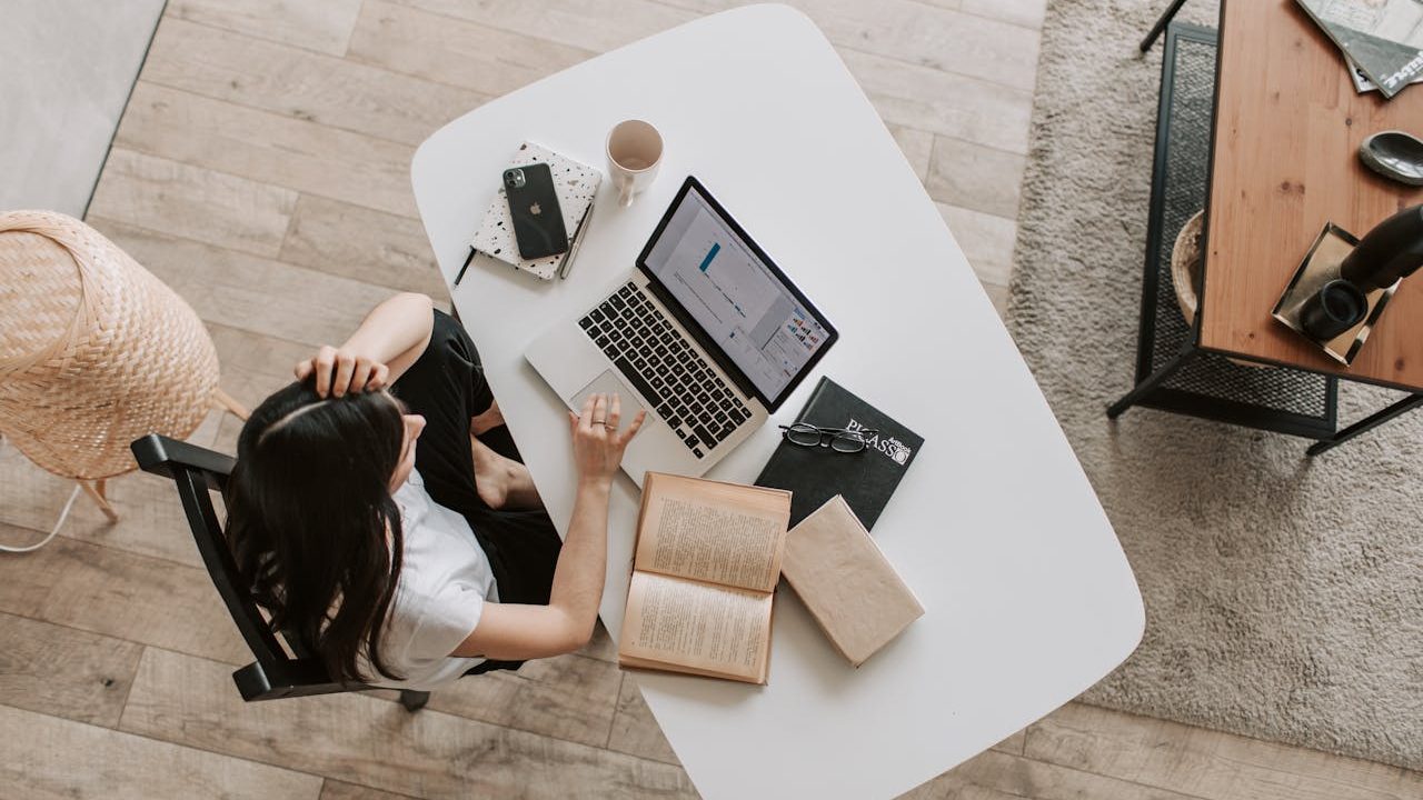 Woman working from home at a white desk with a laptop, books, coffee mug, and phone, viewed from above.