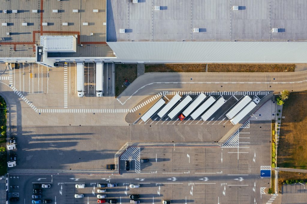 An aerial view of a logistics facility showing semi-trucks at loading docks, parked trailers, and a nearby car parking lot.