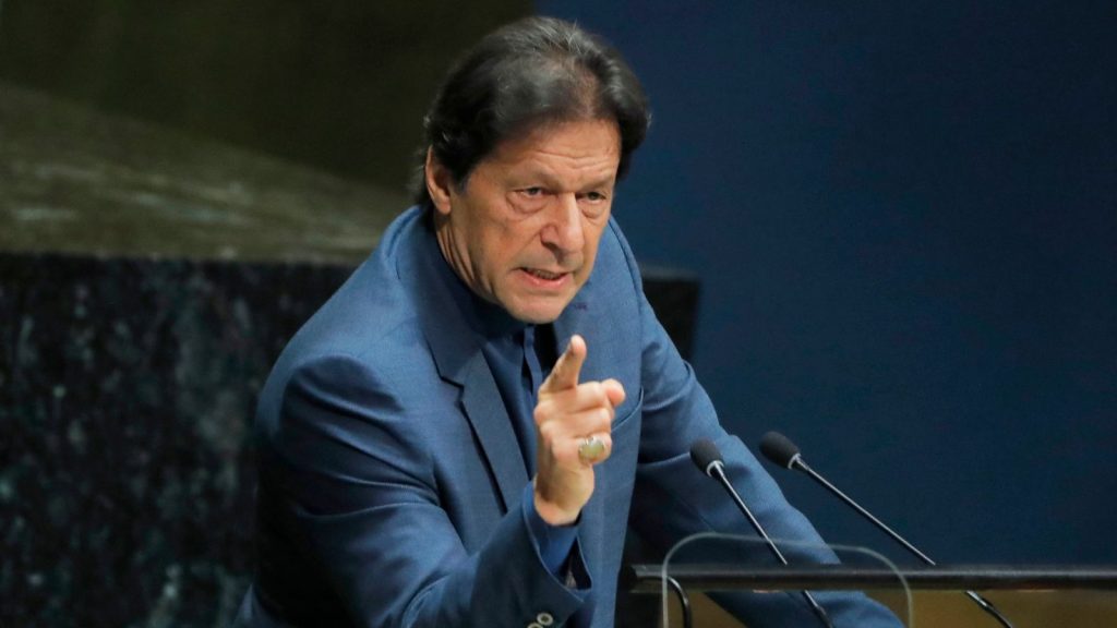 Imran Khan in a blue suit passionately addressing an audience from a podium with multiple microphones, raising his finger in emphasis, conveying urgency and concern in a formal setting.