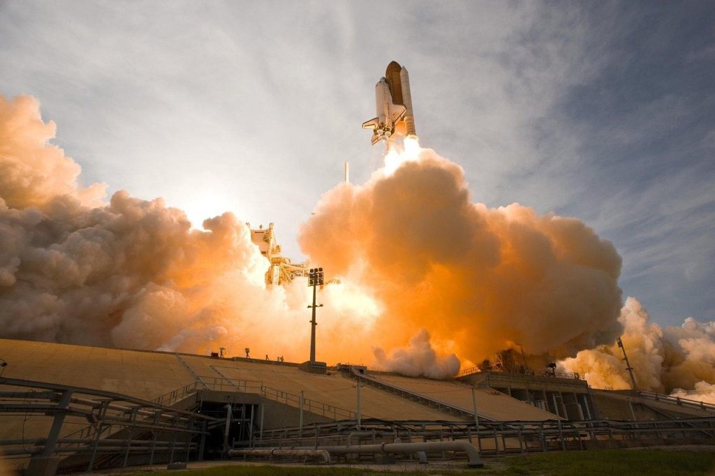 A space shuttle launches from a platform, emitting bright flames and thick clouds of smoke. The shuttle is ascending into the sky with powerful thrust, and the scene is lit dramatically by the sunlight behind the smoke. The launchpad structure and support systems are visible beneath the shuttle.
