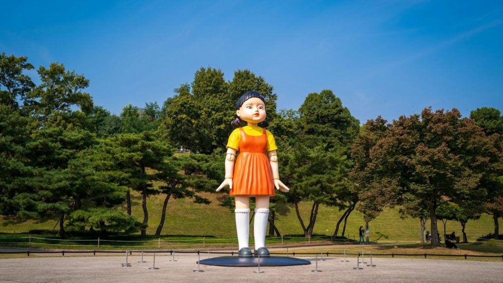 A giant statue of the “Red Light, Green Light” doll from Squid Game stands in an open park area, surrounded by trees and a clear blue sky, symbolizing the show's eerie atmosphere and hinting at its anticipated return in Season 3.