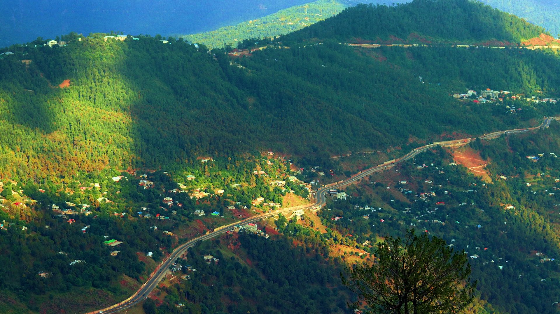 Aerial view of Murree’s lush green hills and winding roads under a cloudy sky, as mysterious weather adds a magical, cloud-blown atmosphere to the landscape.