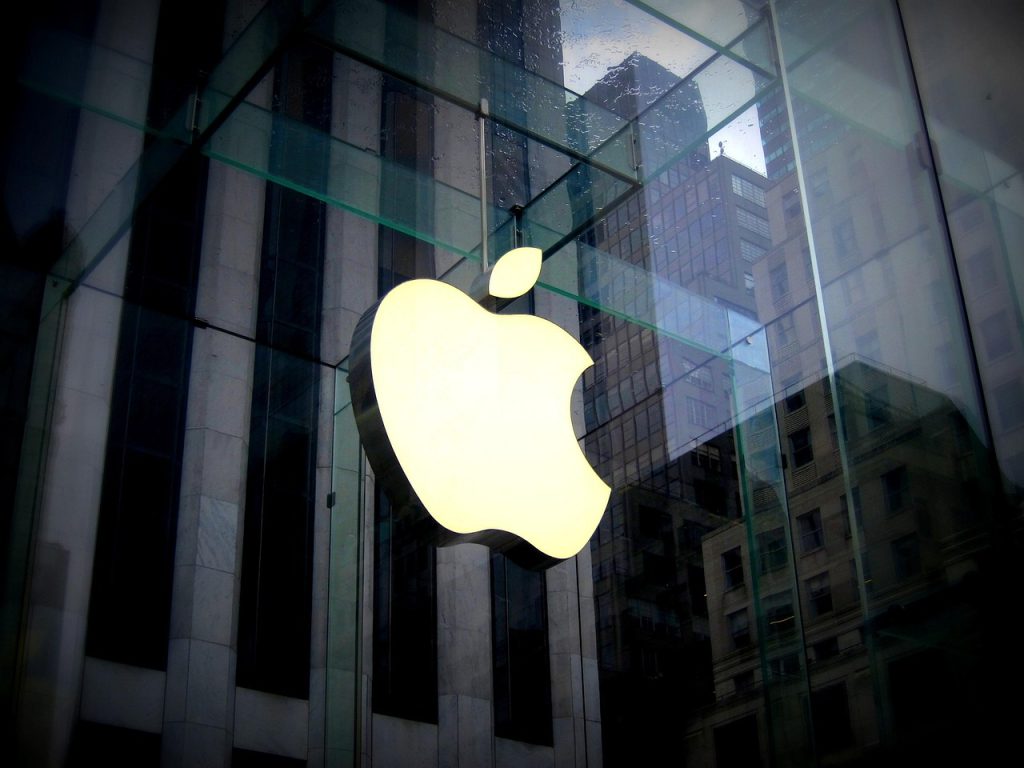 Apple Eyes US F1 Broadcast Rights After Movie Success. A glowing Apple logo displayed on a glass storefront with city buildings reflected in the background.