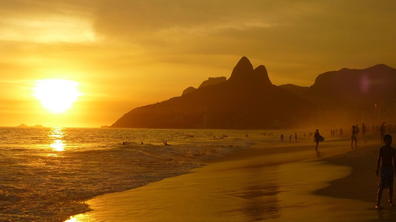 A golden sunset bathes the shoreline in warm hues as people stroll and swim along the historic Hawke’s Bay, with distant rocky hills silhouetted against the sun.
