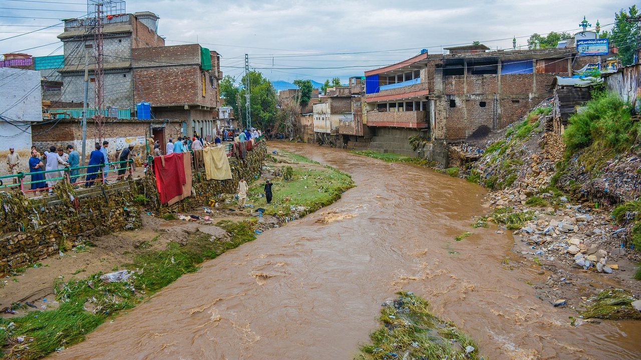 Residents gather along a muddy riverbank in a densely populated area as floodwaters rise under cloudy skies, reflecting NDMA’s monsoon alert for July 6.