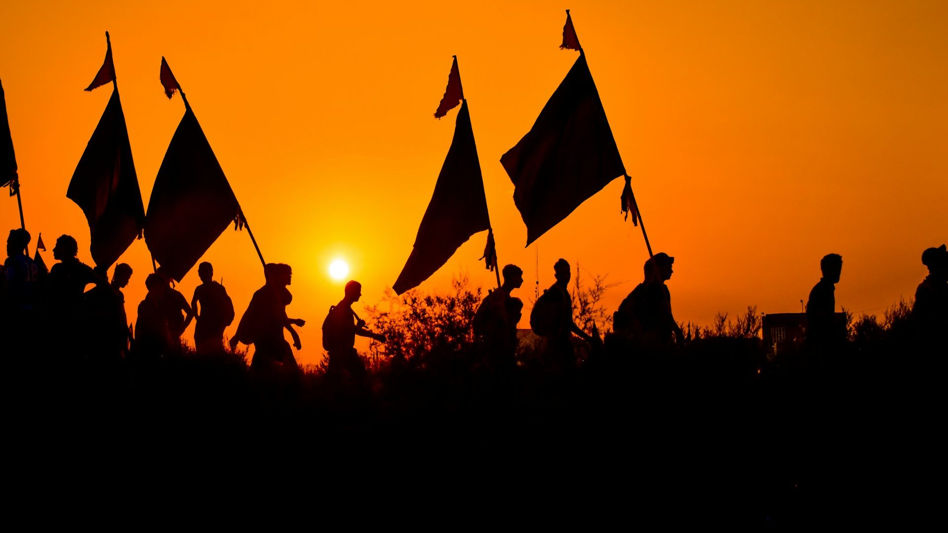Pilgrims carrying flags walk together during sunset on the Arbaeen pilgrimage, symbolizing devotion, with no vehicles or road visible—reflecting the government's ban on road travel for Arbaeen pilgrims.