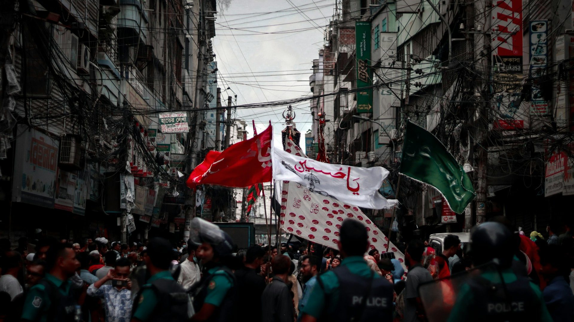 Ashura procession in Karachi amid tight security, with mourners carrying red, white, and green flags through a narrow street lined with buildings and police presence.