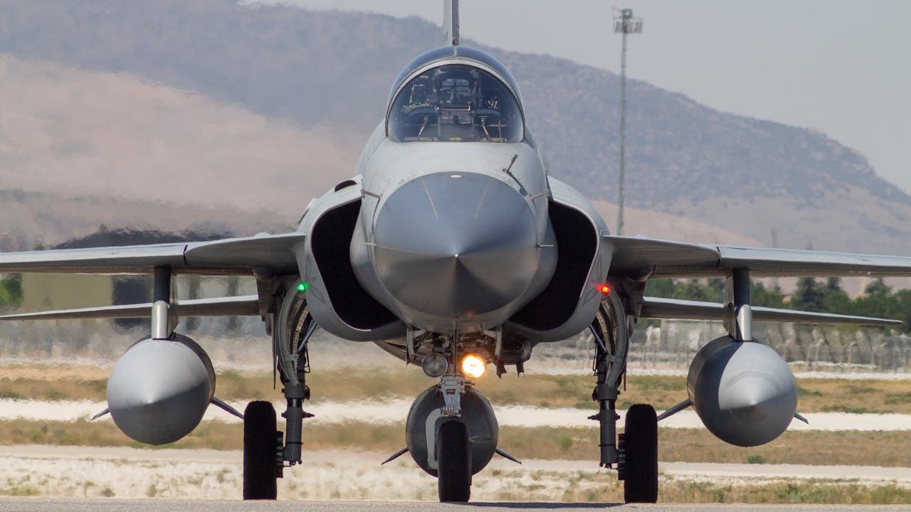 A front-facing view of a Pakistan Air Force JF-17 Thunder fighter jet taxiing on a runway, with mountainous terrain visible in the background.