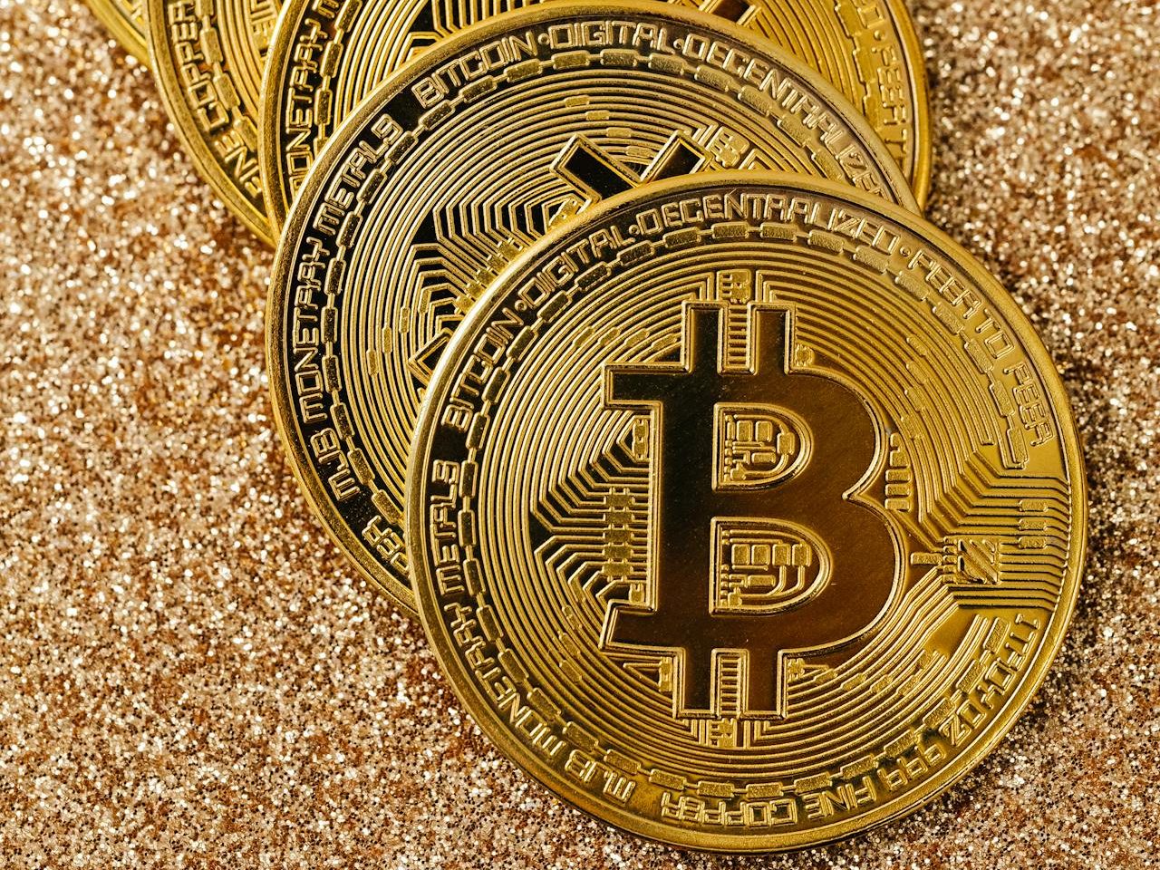 A close-up of five shiny gold-colored Bitcoin tokens arranged in a diagonal line on a glittery gold surface, representing a significant rise in Bitcoin value.