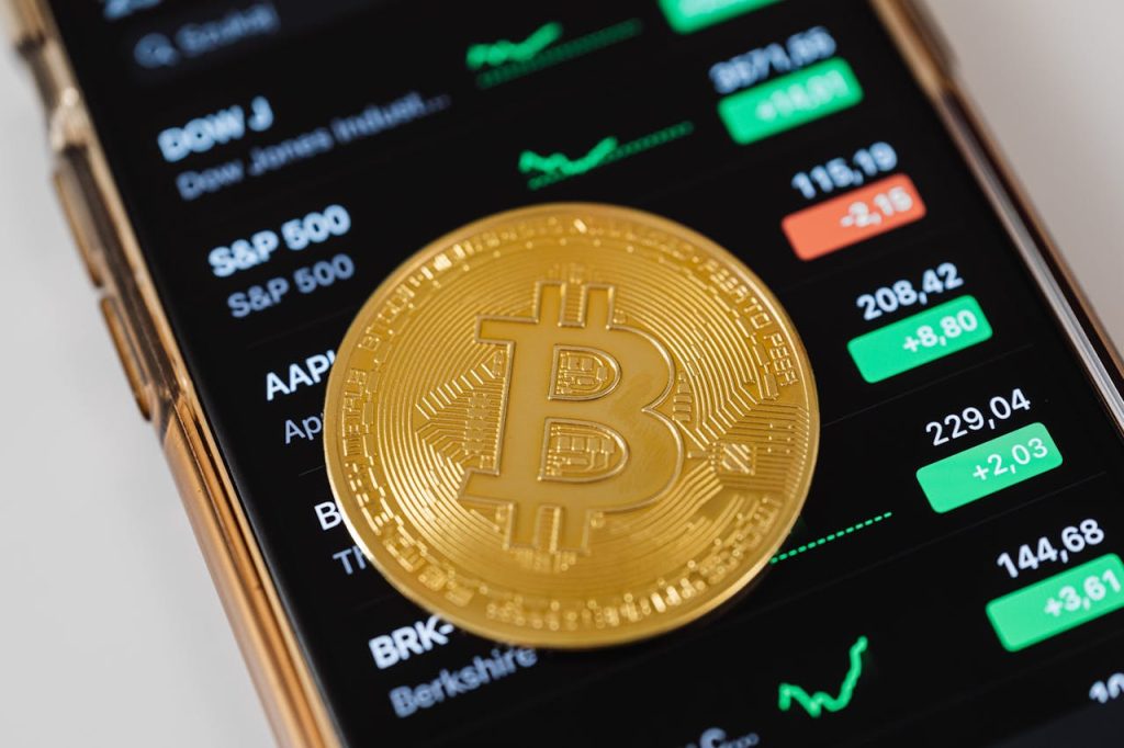 A golden Bitcoin token placed on a smartphone screen displaying stock market data and charts, symbolizing Bitcoin's record-breaking price surge.