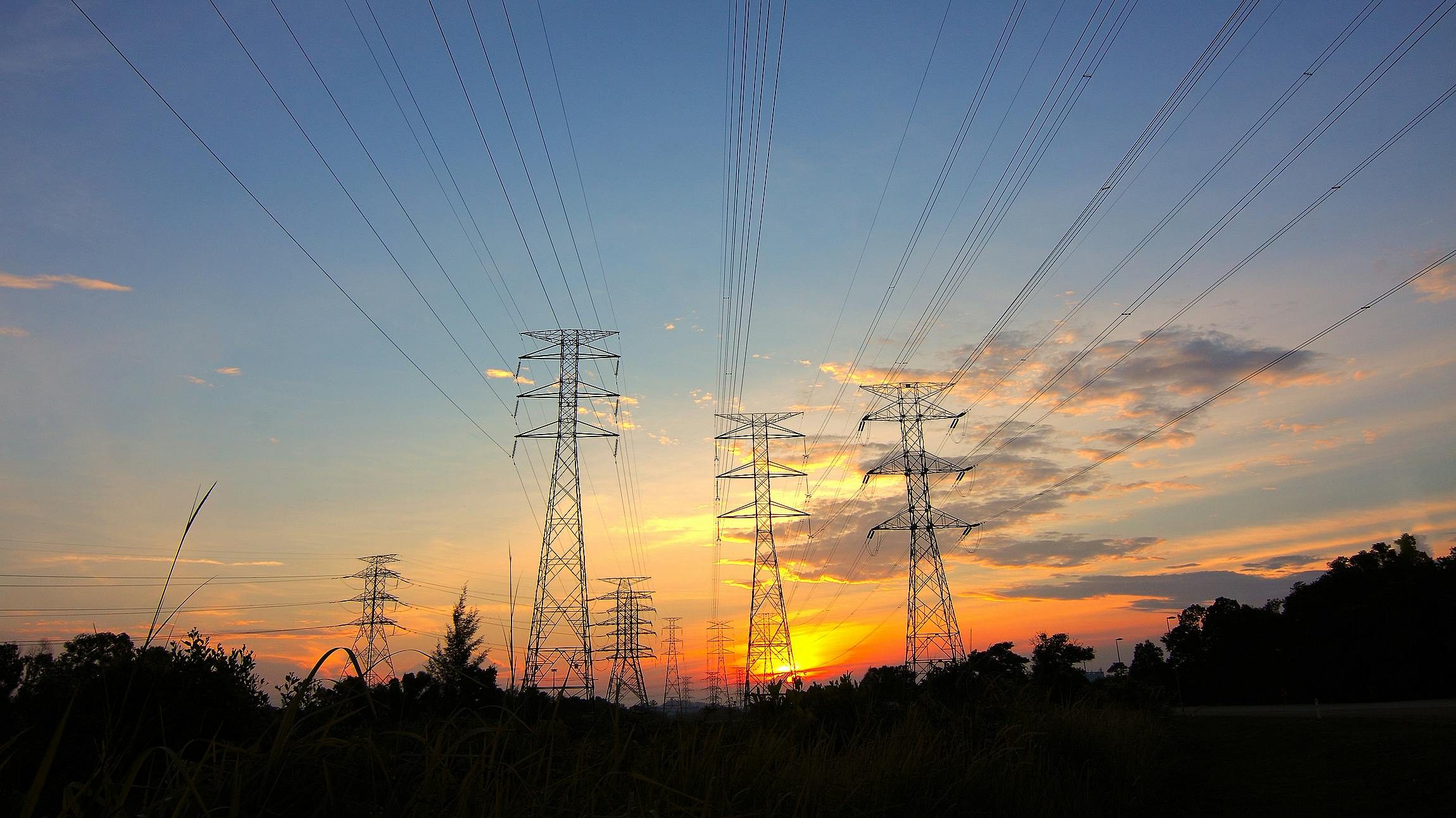 High-voltage power transmission towers stretching into the distance during sunset, symbolizing an underutilized electricity infrastructure amid reduced consumption and rising prices.
