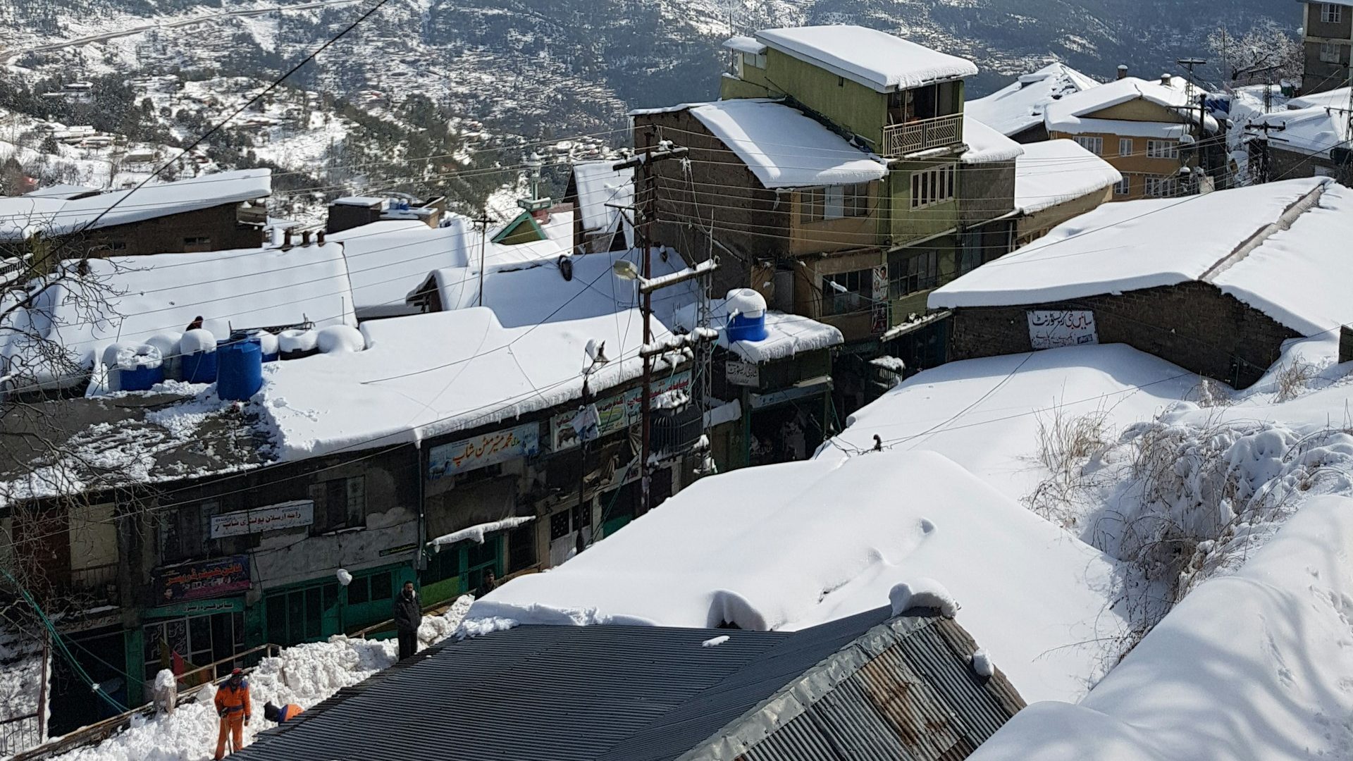 Snow-covered rooftops and buildings in Murree under a partly cloudy sky, with blown clouds drifting over distant mountains — depicting the mysterious weather in this winter paradise.